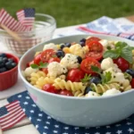A festive red, white, and blue pasta salad with tomatoes, feta cheese, blueberries, and herbs, served in a large bowl on a patriotic-themed 4th of July picnic table.