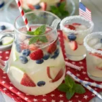 A pitcher of red, white, and blue sangria with sliced strawberries, apple stars, and blueberries in white wine, served in glasses with festive straws on a patriotic 4th of July-themed table.