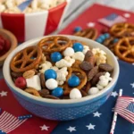 Firecracker trail mix with pretzels, popcorn, dried cranberries, white chocolate chips, and blue candies in a patriotic bowl with 4th of July decorations.