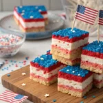 Red, white, and blue layered Rice Krispie treats topped with patriotic sprinkles and Pop Rocks, arranged on a serving tray with festive 4th of July decorations in the background.