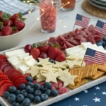 A 4th of July charcuterie board with red, white, and blue fruits, cheeses, meats, and snacks, decorated with American flags and festive party accessories.