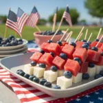 Mini skewers with watermelon cubes, feta cheese, and blueberries arranged on a festive 4th of July platter with patriotic decorations in the background.