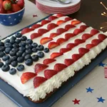 A rectangular flag cake with whipped topping, blueberries in the top-left corner, and rows of strawberries forming red stripes, styled with patriotic decorations for a Fourth of July celebration.