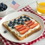 Slice of toast decorated like an American flag with cream cheese, strawberries, and blueberries, served on a white plate with 4th of July decorations.