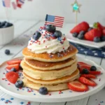 Stack of pancakes topped with whipped cream, strawberries, blueberries, and a mini American flag, served on a festive 4th of July breakfast table.