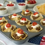 Mini taco cups baked in a muffin tin with ground beef, cheese, and patriotic toppings in red, white, and blue, served on a festive 4th of July picnic table.