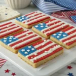Cookie bars decorated like the American flag with red and white frosting stripes, blue frosting in the corner, and white star sprinkles, displayed on a festive 4th of July-themed table.