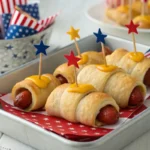Crescent-wrapped Firecracker Hot Dogs standing upright with skewers topped by cheese stars, served on a festive 4th of July platter decorated with patriotic napkins and American flags.