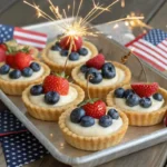Mini fruit tarts with golden crusts, cream filling, and red and blue berries arranged on a tray with patriotic 4th of July decorations.