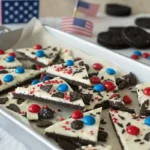 White chocolate Oreo bark with red and blue M&Ms, crushed Oreos, and patriotic sprinkles, broken into pieces on a tray with 4th of July decorations.