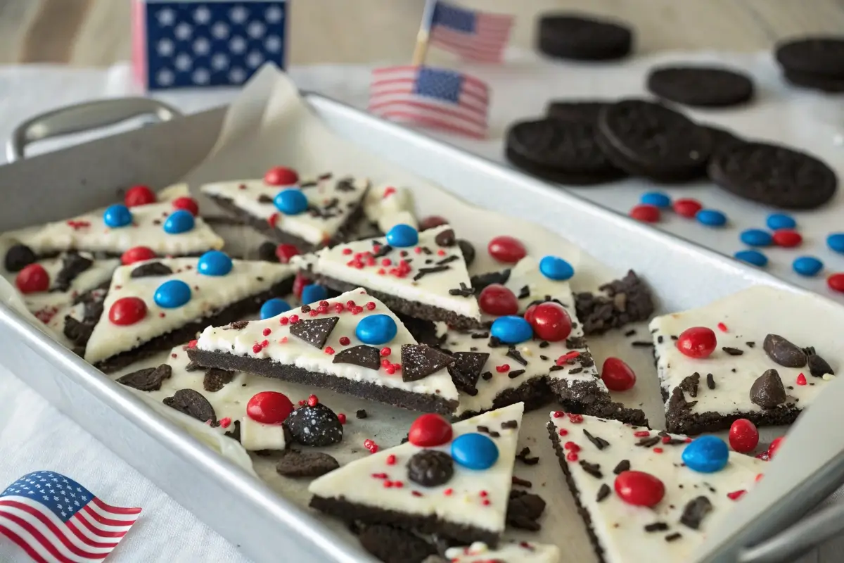 White chocolate Oreo bark with red and blue M&Ms, crushed Oreos, and patriotic sprinkles, broken into pieces on a tray with 4th of July decorations.