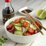 Cucumber and tomato salad with sesame seeds and soy dressing, served in a ceramic bowl with chopsticks and lime wedges on a light wood surface.