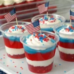 Individual clear cups layered with red, white, and blue Jello, topped with whipped cream and patriotic sprinkles, decorated with small American flags and set on a festive Fourth of July table.
