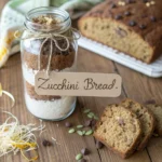 Mason jar layered with dry zucchini bread ingredients, ribbon, and recipe card tag attached.