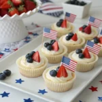 Mini cheesecakes decorated to look like American flags with blueberries, sliced strawberries, and whipped cream, displayed on a patriotic-themed tray for a 4th of July celebration.