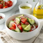 Healthy cucumber tomato salad without oil in a white ceramic bowl, with lemon-vinegar dressing and red onion slices on a light table.