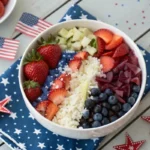 Red, white, and blue rice bowl with tomatoes, blueberries, feta, and herbs arranged in a patriotic design for 4th of July, served in a white bowl with festive decorations.