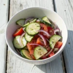 Overhead photo of cucumber tomato salad in a ceramic bowl, with sliced red onions and a glossy olive oil and vinegar dressing on a rustic wooden table.