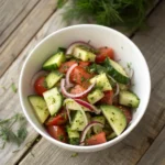Bowl of chopped cucumbers and tomatoes with thin red onions and fresh herbs, lightly dressed, on a rustic wooden table with natural lighting.