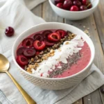 Top-down macro shot of a cherry smoothie bowl with toppings arranged in neat rows: cherries, seeds, coconut flakes, and granola.
